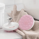 A pink and white pet grooming brush with a textured pink silicone scrubber head and a white handle, displayed next to a beige towel and a white bottle in a bathroom setting.