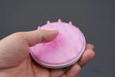 A hand holding a small pink scalp massager brush with soft silicone bristles and a white base, shown against a dark gray background.