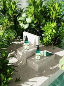 Three green and white skincare bottles from the brand C-TO with black pumps are arranged on a light gray stone platform surrounded by lush green plants in a sunny outdoor setting.