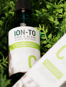 A dark glass bottle labeled ION-TO Hair & Scalp Science Specialist, placed among green leaves with part of a white and green product box visible in the foreground.