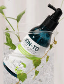 A dark blue pump bottle labeled 'ION-TO Hair & Scalp Silence Specialist' resting inside a clear glass beaker filled with ice, with green leaves draped around it against a light neutral background.