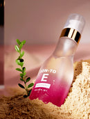 A frosted glass bottle of EON-TO E Hair Essence with a pink-to-clear gradient and a gold collar, partially embedded in sand next to a small green plant sprig.