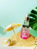 Pink and clear bottle of ION-TO E Hair Essence styled in a beach scene with sand, a small yellow umbrella, a striped beach ball, and green leaves against a mint green background.