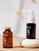 Two amber glass hair tonic bottles with white caps and pump tops, labeled NON-TO XENON & SCALP G, displayed on a light wood surface with a white ceramic dish.