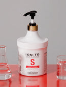 A white plastic pump bottle of HISTEMO ION-TO Hair & Scalp Science Specialist Scalp Cleaner with a gold collar and black pump, set on a reflective red surface with two glass beakers on either side against a gray background.