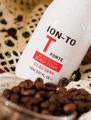 A white bottle of ION-TO T Forte Scalp Tonic surrounded by coffee beans on a textured beige surface.
